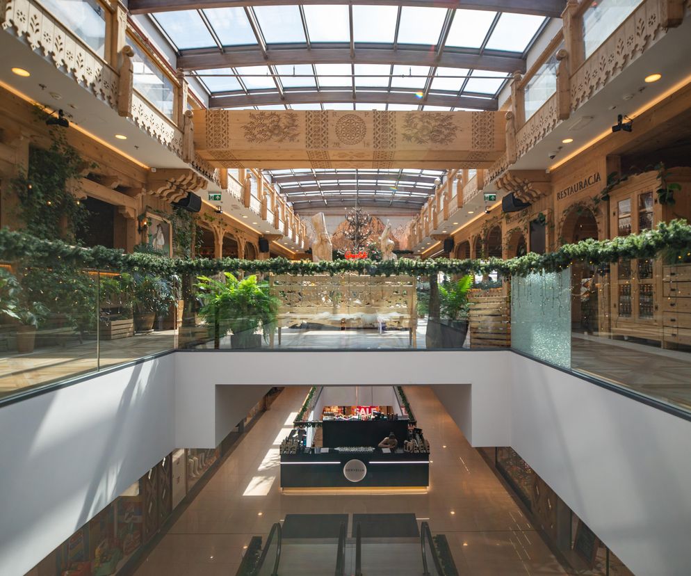 Wnętrze centrum handlowego Krupówki 40 w Zakopanem. Widoczne piętra z balustradami, witrynami sklepów, oraz drewnianymi elementami dekoracyjnymi w stylu zakopiańskim. Na górze szklany dach przepuszczający światło. Aranżacje wnętrz i architekturę obiektów na Krupówkach omawia Architektura Murator Plus.