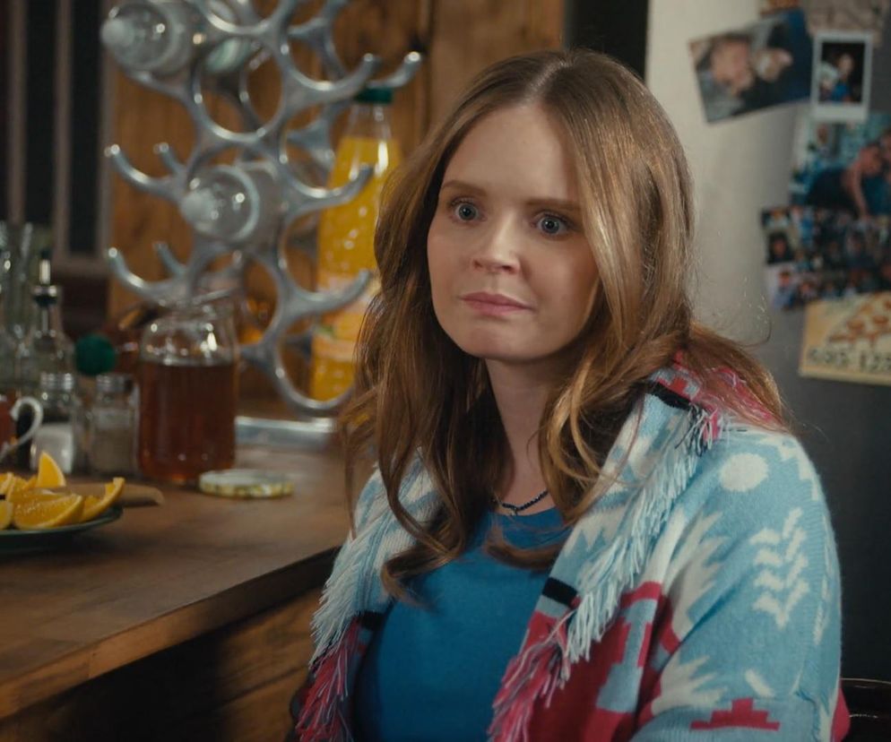 Magda (Olga Kalicka) z serialu Rodzinka.pl siedzi przy drewnianym stole w kuchni, ubrana w błękitną bluzkę i wzorzysty sweter. Obok niej kawałki pomarańczy i naczynia. W tle lodówka z magnesami. O dramacie Magdy dowiesz się więcej na portalu Super Seriale.