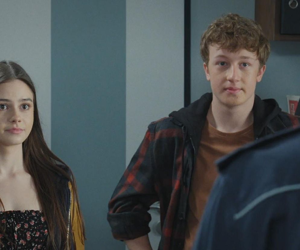 Basia (Karina Woźniak) i Kacper (Bartosz Ziewiec) w rozmowie z policjantem, na zdjęciu Basia w żółtej kurtce z frędzlami, a Kacper w koszuli w kratę. Szukają sprawcy wypadku Franki i Pawła, a o tym, jak przebiega śledztwo, przeczytasz na Super Seriale.