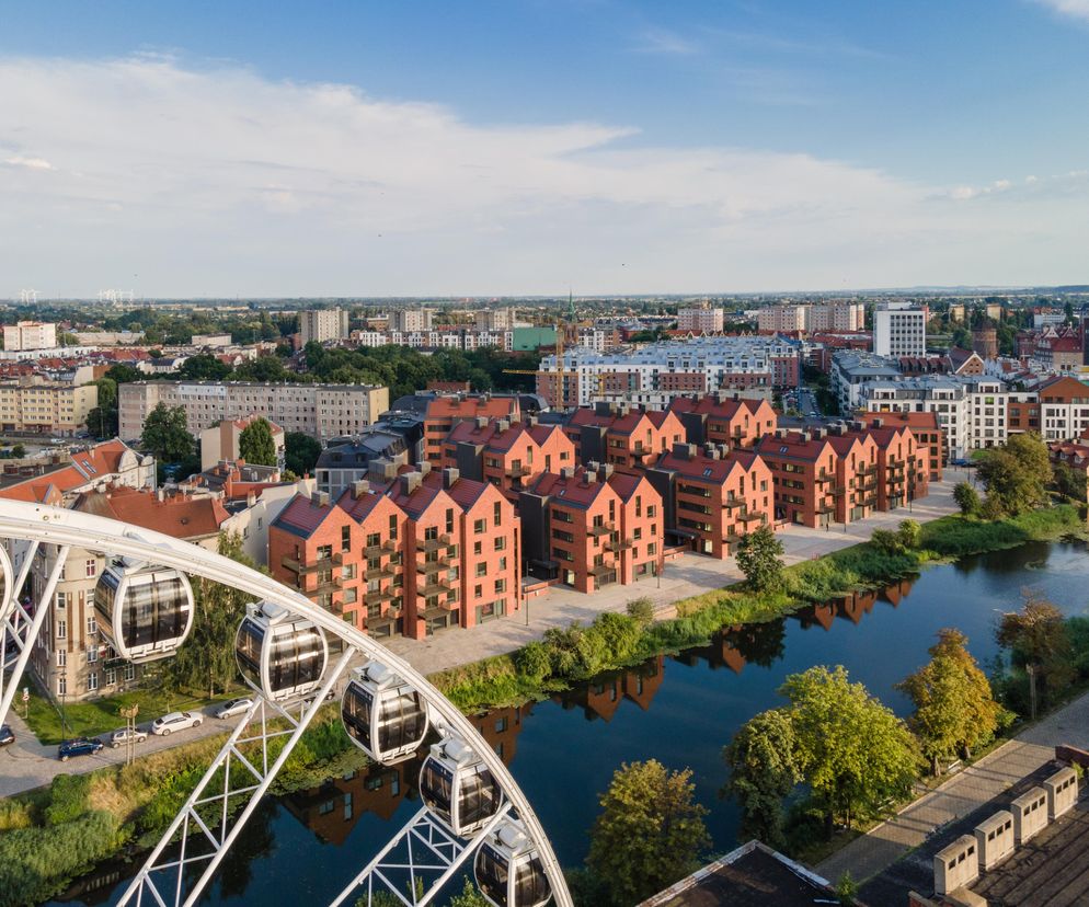 Z lotu ptaka widać Riverview w Gdańsku, zespół nowoczesnych, ceglanych budynków o spadzistych dachach, otoczony zielenią i rzeką, z odbiciem elewacji w wodzie. Na pierwszym planie fragment diabelskiego młyna. Więcej o tym wyjątkowym projekcie autorstwa Szymona Wojciechowskiego na Architektura Murator Plus.