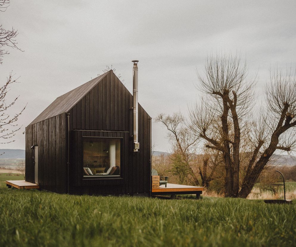 Ciemna bryła domu Hyttee 2.0 z drewnianą elewacją z modrzewia, osadzona na zielonym trawiastym terenie z widocznym tarasem i oknem, które otwiera wnętrze. Projekt zgłoszony do konkursu ŻYCIE W ARCHITEKTURZE 2025, o którym przeczytasz więcej na Architektura Murator Plus.