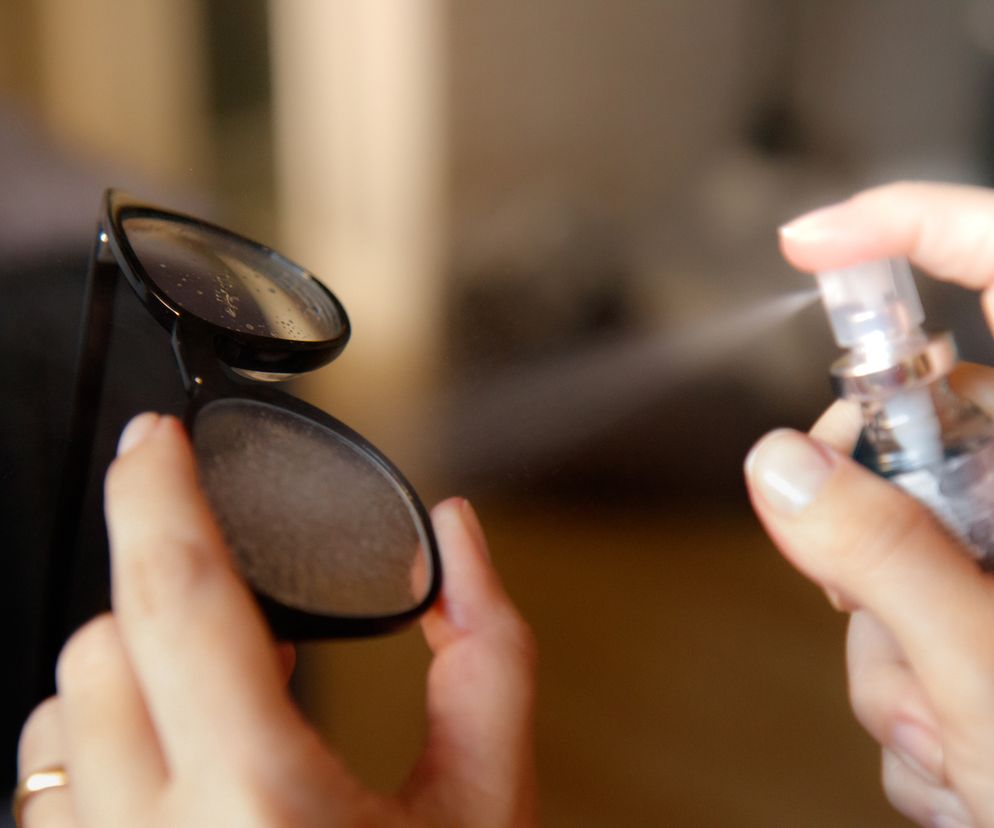 Kobieta spryskuje soczewki okularów korekcyjnych preparatem zapobiegającym parowaniu. Okulary z czarnymi oprawkami są trzymane w lewej dłoni z pierścionkiem, a prawa dłoń rozpyla płyn. To skuteczne sposoby na zaparowane okulary w maseczce. Więcej informacji znajdziesz na Poradnik Zdrowie.