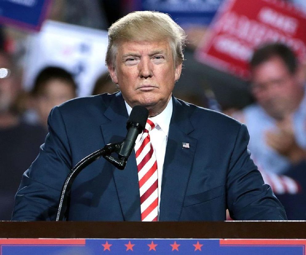 Donald Trump w granatowym garniturze i krawacie w czerwono-białe pasy, stojący za mównicą z mikrofonem. Prezydent USA ma poważny wyraz twarzy, jego wypowiedzi na temat paracetamolu i autyzmu wywołują debatę, o czym można przeczytać na Poradnik Zdrowie.