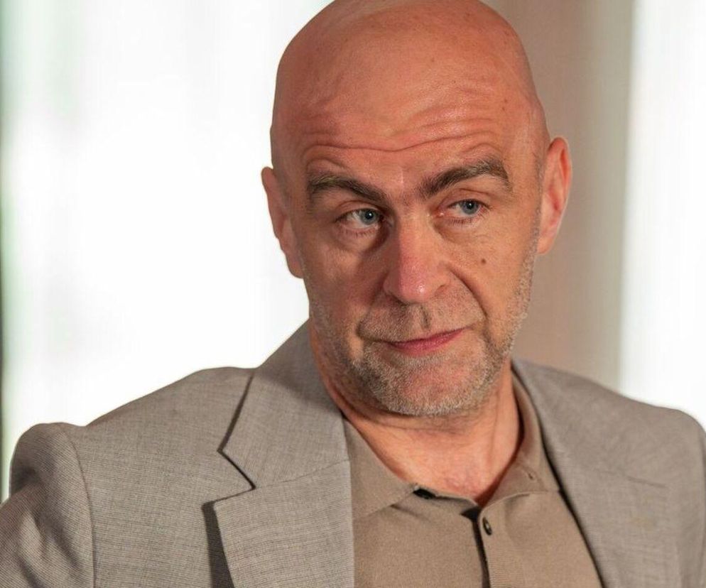 Łysy, brodaty mężczyzna, Bazylow z serialu Barwy Szczęścia, w szarym garniturze i beżowej koszulce, patrzy w bok z poważną miną. Scena odzwierciedla kontekst, w którym Bazylow grozi Stefaniakowi. Więcej o losach bohaterów przeczytasz na Super Seriale.