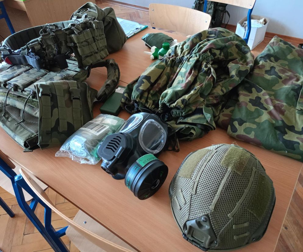 Wyposażenie wojskowe, w tym hełm, maska gazowa i mundur na stole. O Wakacjach z Wojskiem przeczytasz na Eska Siedlce.