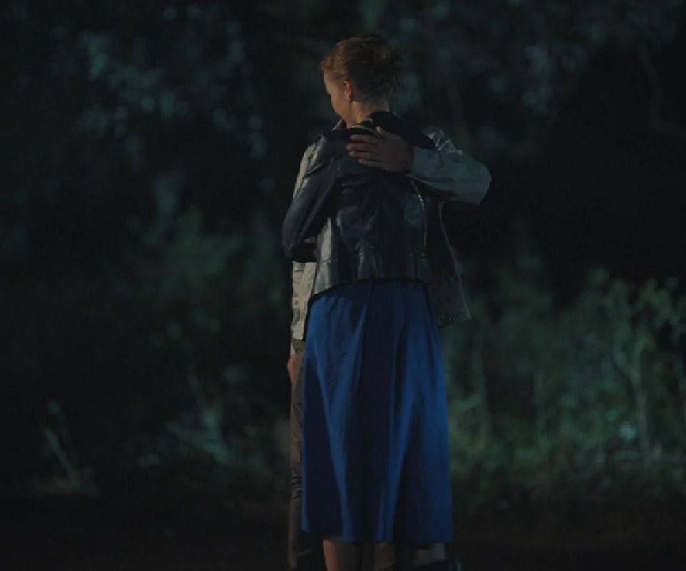 Para, Natalka (Dominika Suchecka) i Adam (Patryk Szwichtenberg) z serialu M jak miłość, obejmuje się na tle ciemnych drzew w nocnej scenerii. Kobieta ubrana jest w niebieską spódnicę i ciemną kurtkę. Scena przedstawia romantyczne zbliżenie bohaterów. Więcej o ich losach znajdziesz na Super Seriale.