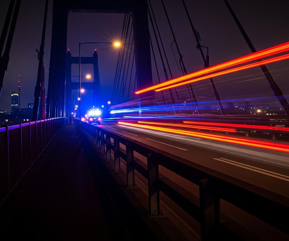 Nocny widok z mostu z rozmazanymi smugami świateł pojazdów w ruchu. Po lewej stronie dominuje ciemna konstrukcja mostu, z której zwisają liny, a w oddali widać podświetlone wieżowce miasta. Po prawej stronie biegnie dwukierunkowa droga z białymi liniami, na której intensywne czerwone i białe smugi światła oznaczają pojazdy oddalające się, natomiast niebieskie smugi symbolizują światła pojazdów nadjeżdżających. Latarnie uliczne emitują pomarańczowe światło, a barierki mostu po lewej stronie mają fioletowe podświetlenie.