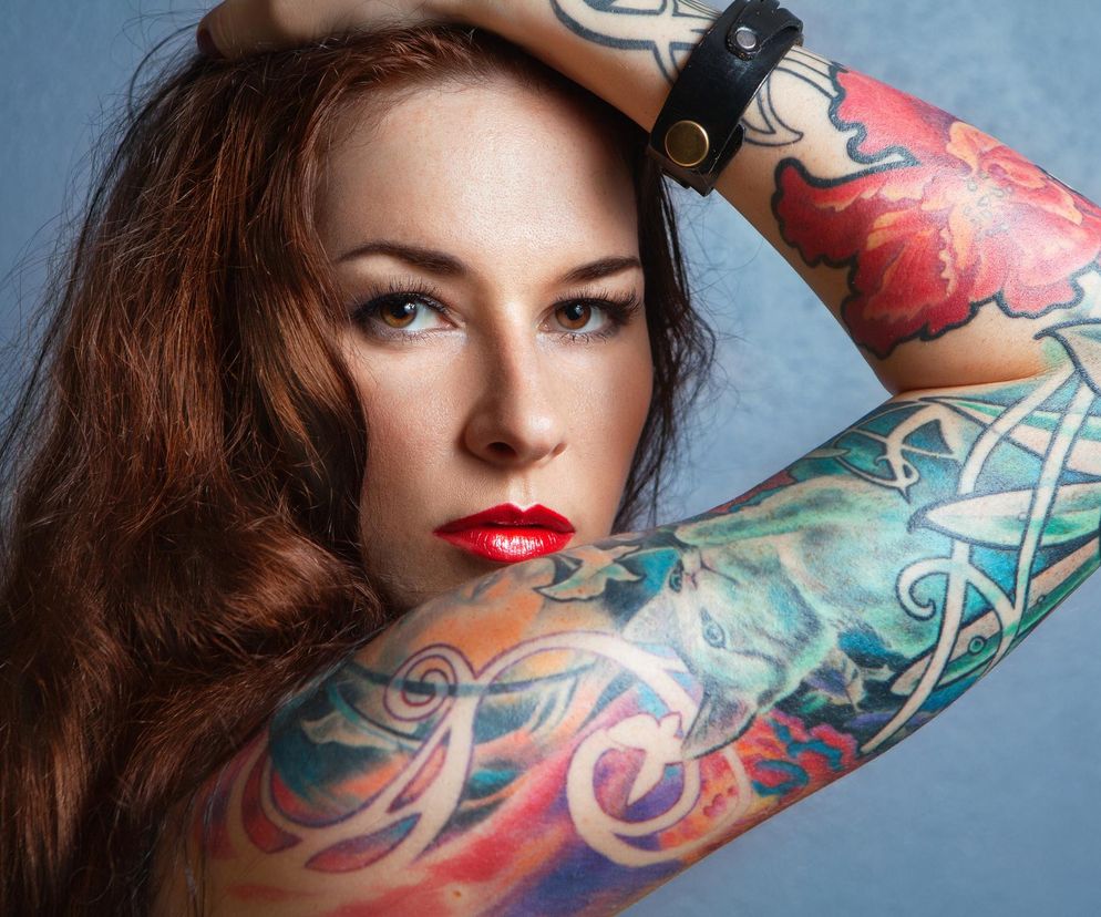 Kobieta z długimi, brązowymi włosami i licznymi, kolorowymi tatuażami na obu ramionach, pozująca na jasnoniebieskim tle. Ma intensywny makijaż i pomalowane na czerwono usta, a dłoń uniesioną do głowy. Na jej ramieniu widoczne są wzory kwiatowe, zwierzęce i celtyckie. Na temat badań o tatuażach i czerniaku przeczytasz na Poradnik Zdrowie.