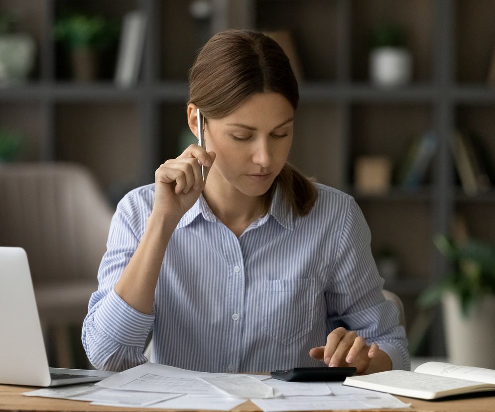 Kobieta w koszuli w paski, skupiona, obliczająca coś na kalkulatorze przy biurku z laptopem i dokumentami. Od 2026 r. KFS otworzy się na samozatrudnionych. Więcej o rewolucji w dofinansowaniach przeczytasz na Super Biznes.