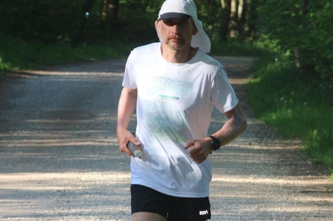 Ultramaratończyk Łukasz Wróbel Motyla Noga biegnący leśną ścieżką w czapce z daszkiem i białej koszulce. W tle widać zieleń drzew. Opis biegu Wróbla i jego celu charytatywnego dla Fundacji Na Ratunek Dzieciom z Chorobą Nowotworową znajdziesz na naszym portalu.