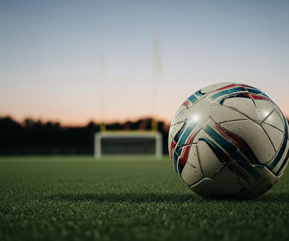 Piłka do piłki nożnej, częściowo biała z czerwonymi i niebieskimi pasami, leży na zielonej trawie stadionu sportowego. W tle, pod wieczornym niebem przechodzącym od jasnego błękitu do pomarańczowego, widoczne są dwie rozmyte, świecące latarnie stadionu oraz ciemne sylwetki drzew i bramka.