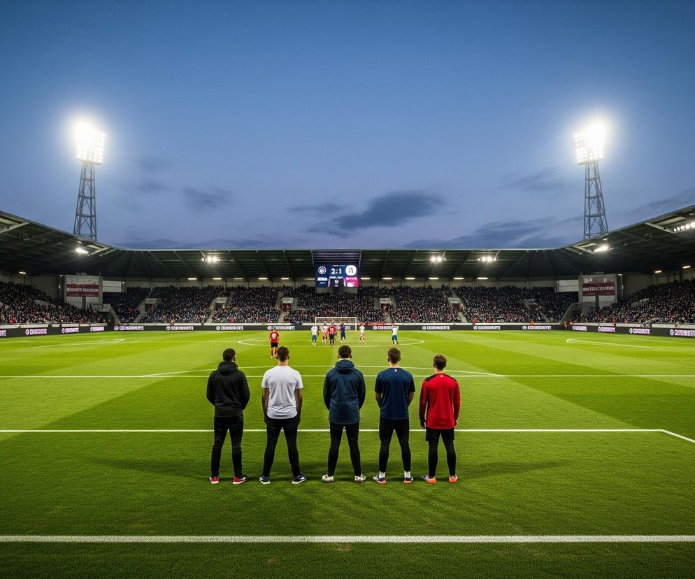 Pięciu mężczyzn w ubraniach sportowych stoi tyłem do widza, obserwując boisko do piłki nożnej oświetlone jaskrawymi reflektorami. Na tle wyraźnie zielonej trawy stadionu z białymi liniami, widoczne są trybuny wypełnione kibicami, a także drużyny piłkarskie w trakcie meczu. Na środku, nad boiskiem, umieszczona jest tablica wyników wyświetlająca rezultat 2:1.