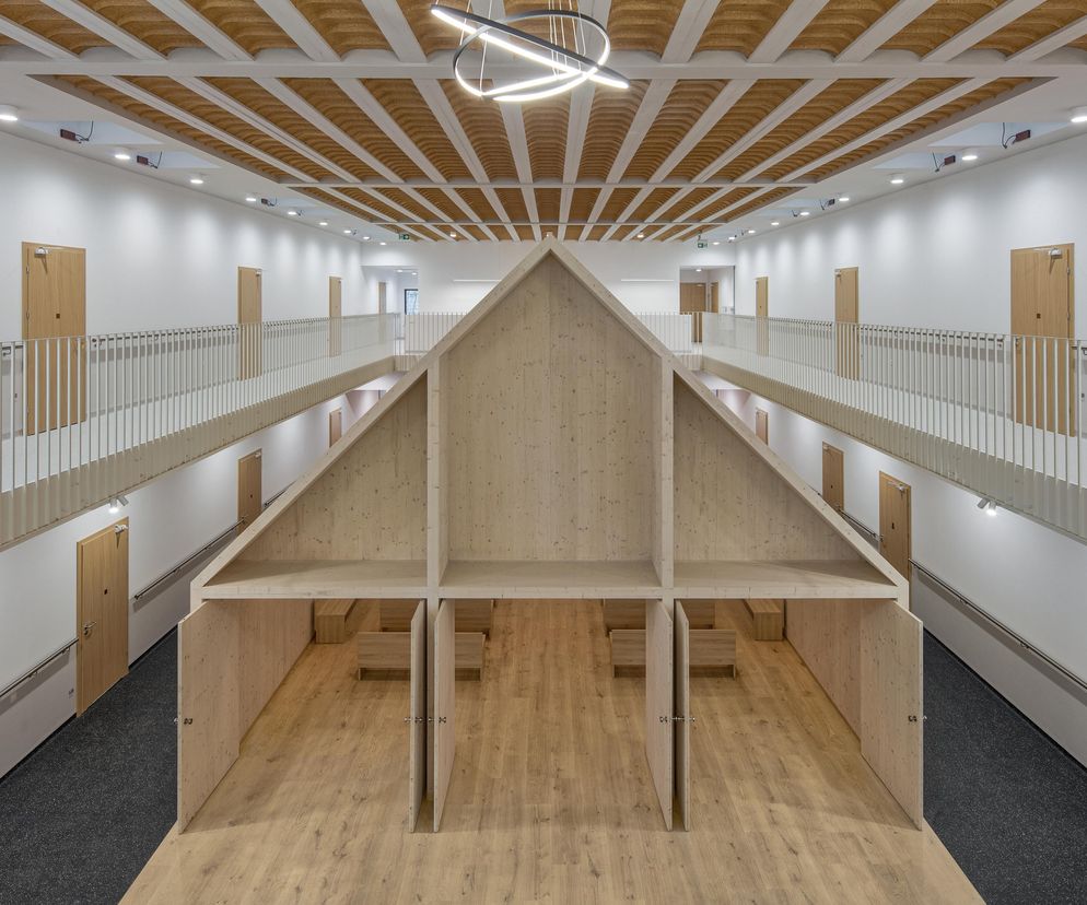 Wnętrze Domu Matki Bożej Serdecznej, widziane z góry, z centralnie umieszczoną konstrukcją z jasnego drewna imitującą kształt domu, otoczoną korytarzami z jasnymi ścianami, drewnianymi drzwiami i białymi balustradami. Więcej o architekturze społecznej na Architektura Murator Plus.