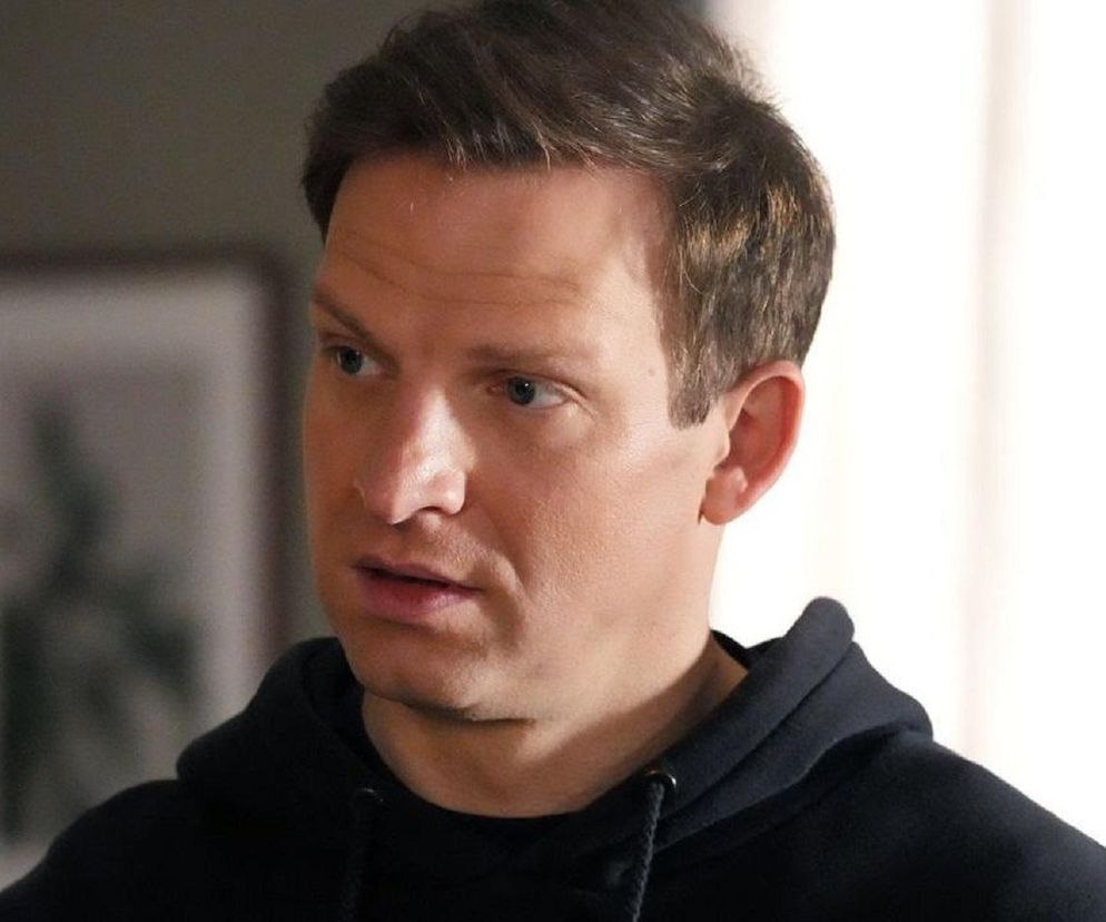 Mężczyzna, aktor Michał Rolnicki w roli Łukasza z serialu Barwy szczęścia, ubrany w czarną bluzę z kapturem, z zaniepokojoną miną. Zdjęcie ilustruje fragment fabuły, w którym Agnieszka przestaje odpowiadać na jego wiadomości. Więcej szczegółów na Super Seriale.