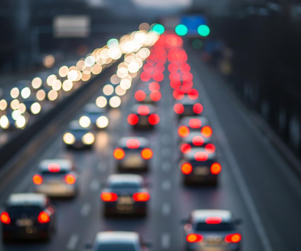 Autostradę w nocy, z widokiem skierowanym lekko w dół, pokazującym rozmyte światła samochodów. Na lewo znajduje się jeden pas ruchu z rzędem żółtawych świateł przednich, które stają się coraz jaśniejsze w miarę oddalania się w głąb obrazu. Na prawo widoczne są dwa pasy ruchu, na których czerwone światła tylne samochodów tworzą dwie kolumny jasnych, rozmytych punktów, również zanikających w oddali. Cała scena jest zamglona i rozmyta, z ciemnym otoczeniem, jedynie światła są wyraźne.