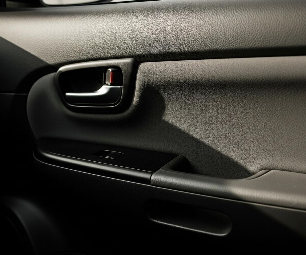 Centralną część obrazu zajmuje ciemnoszara tapicerka drzwi samochodowych o fakturze drobnych ziarenek, rozświetlona ukośnym pasmem światła przechodzącym przez jej górną część. Pośrodku znajduje się czarna klamka wewnętrzna z chromowanym uchwytem i wyraźnym czerwonym wskaźnikiem blokady drzwi. Poniżej klamki umieszczony jest czarny panel z przełącznikiem do sterowania szybą, a w lewym górnym rogu widać fragment kratki nawiewu powietrza z metalicznym obramowaniem. Dolna część kadru jest znacznie ciemniejsza, prawie czarna, z delikatnie widocznym wgłębieniem uchwytu na rękę i innymi subtelnymi detalami struktury.