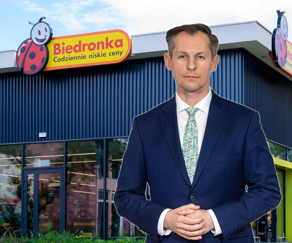 Prezes UOKiK Tomasz Chróstny na tle sklepu Biedronka. Stoi w garniturze i krawacie. Zdjęcie ilustruje zarzuty UOKiK wobec sieci Biedronka. Więcej na ten temat przeczytasz na portalu Super Biznes.