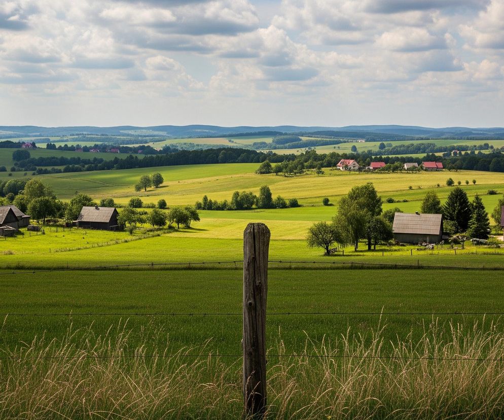 Drewniany słup ogrodzeniowy znajduje się na pierwszym planie, w centralnej części zdjęcia, przed pasem suchej trawy i drutami kolczastymi rozciągającymi się horyzontalnie. Za nim rozciąga się zielone pole, a w tle widać malowniczy wiejski krajobraz z licznymi domami o czerwonych i szarych dachach, rozsianymi wśród drzew i kolejnych pól. Na horyzoncie widoczne są pofałdowane wzgórza, a nad całością rozciąga się niebo pokryte białymi, kłębiastymi chmurami.