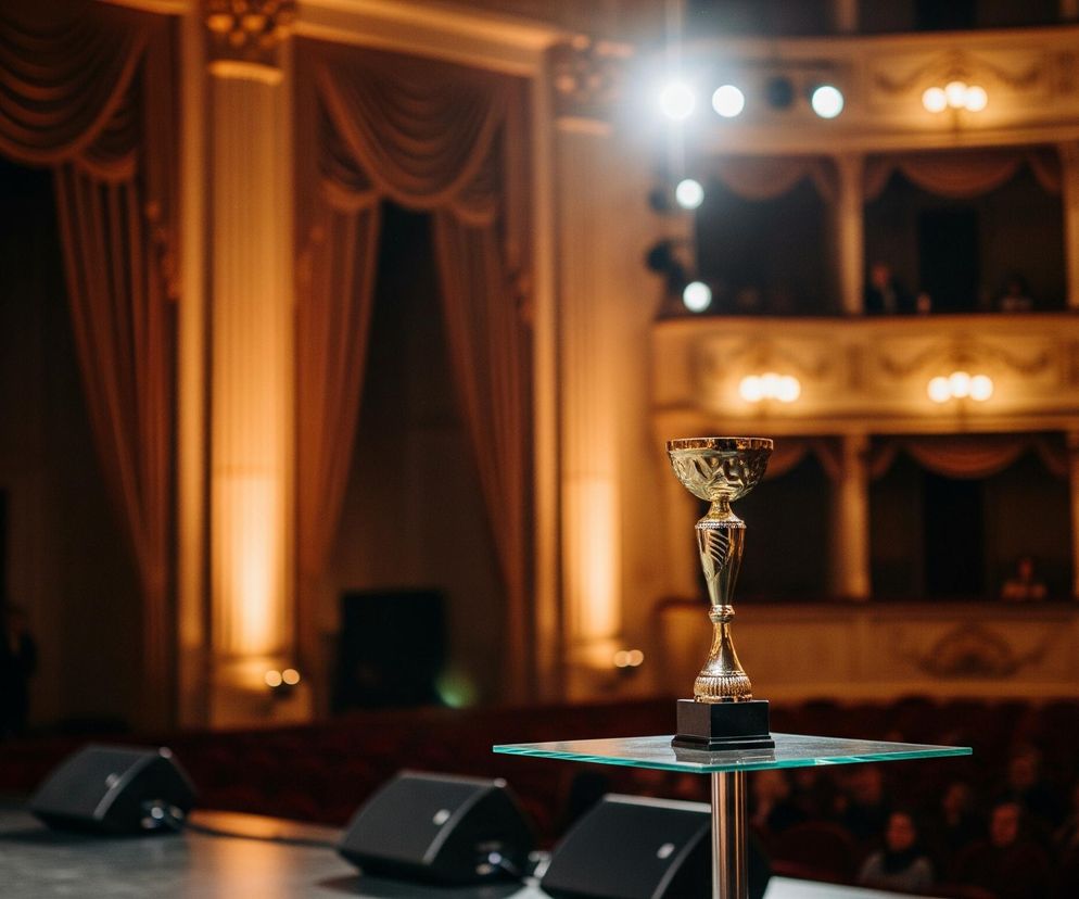Złoty puchar stoi na szklanym blacie małego stolika na scenie, oświetlony jasnym światłem reflektora z góry. Scena jest ciemna, a w tle widać ciemne, pomarańczowo-brązowe zasłony oraz puste loże teatru z nielicznymi, rozmazanymi postaciami. Obok stolika z pucharem leżą czarne głośniki, a po lewej stronie znajduje się drugi, mniejszy stolik z niewyraźnym złotym pucharem.