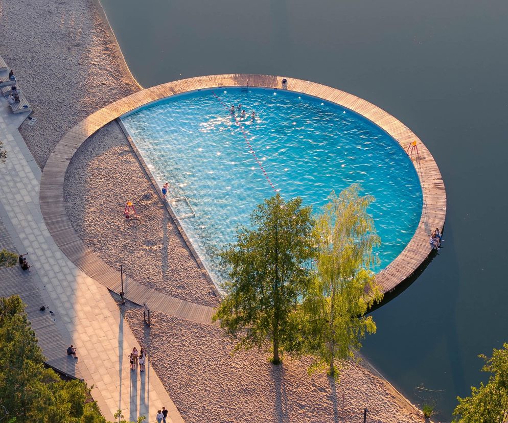 Widok z lotu ptaka na okrągły, błękitny basen na jeziorze, otoczony drewnianym pomostem i piaszczystą plażą. Na basenie widać kąpiących się ludzi, a na plaży i promenadzie spacerowiczów i osoby odpoczywające na ławkach. Po lewej stronie bujna roślinność, w tle ciemna woda jeziora. Projekt zagospodarowania terenów rekreacyjnych, o którym przeczytasz więcej na Architektura Murator Plus.