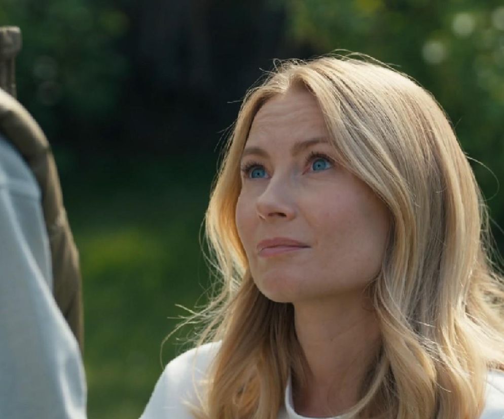 Uśmiechnięta Karolina Chapko jako Dominika z serialu Barwy szczęścia w białej bluzce z dekoltem, z długimi, blond włosami i niebieskimi oczami, spogląda w prawą stronę na Mateuszka, który odwrócony stoi tyłem. Aktorka jest gotowa prosić o przebaczenie w imieniu swoim i Sebastiana, o czym przeczytasz więcej na Super Seriale.