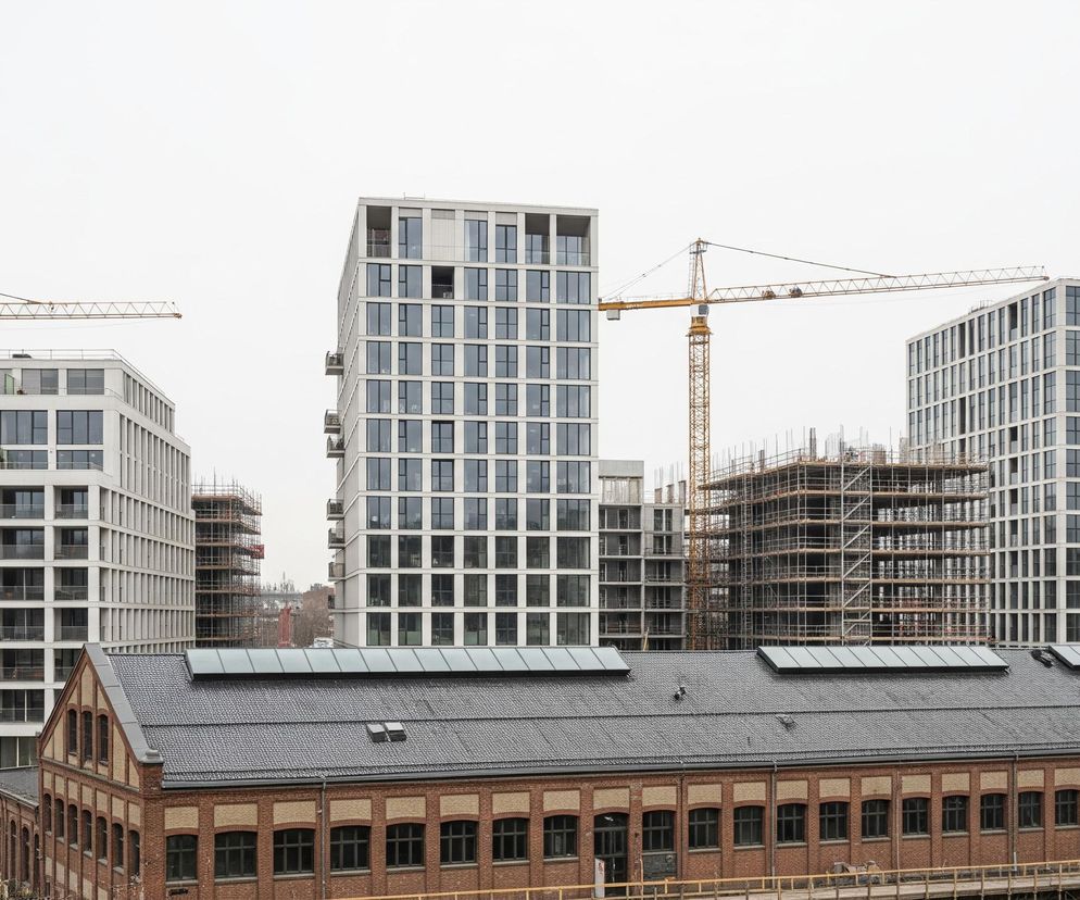 Centralnym elementem obrazu jest duży, ceglany budynek z ciemnym, spadzistym dachem, pokrytym dachówką, z widocznymi świetlikami. Przed budynkiem, na pierwszym planie, biegnie drewniana konstrukcja przypominająca most lub podest. Za ceglanym budynkiem wznoszą się nowoczesne, białe budynki mieszkalne z licznymi oknami i balkonami, część z nich jest jeszcze w fazie budowy, otoczona rusztowaniami i żółtymi żurawiami budowlanymi. Tło stanowi jasne, pochmurne niebo.