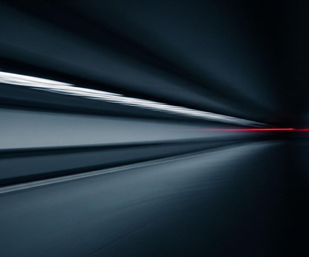 Centralnie umiejscowiony, rozciągnięty, intensywnie czerwony pasek światła przecina niemal cały kadr, tworząc silny punkt ostrości. Otacza go dynamiczna kompozycja, gdzie dominuje czerń i ciemne odcienie szarości, rozmyte, tworzące wrażenie szybkiego ruchu i tunelowego efektu. Po lewej stronie, wyżej od czerwonej linii, widoczne są jaśniejsze, rozmazane białe smugi, równoległe do siebie i skierowane w głąb obrazu. Dolna część kadru jest nieco jaśniejsza, z subtelnymi odcieniami szarości, również z efektem rozmycia.