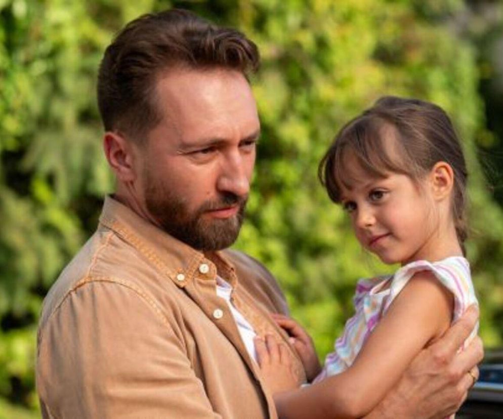 Piotr Strzelecki trzyma w ramionach córkę Lidzię, z zamyśloną miną, patrząc w bok. Dziewczynka w jasnej sukience opiera się o ojca, patrząc z zaciekawieniem w kierunku widza. Zdjęcie z serialu Na sygnale dostępne na Super Seriale.