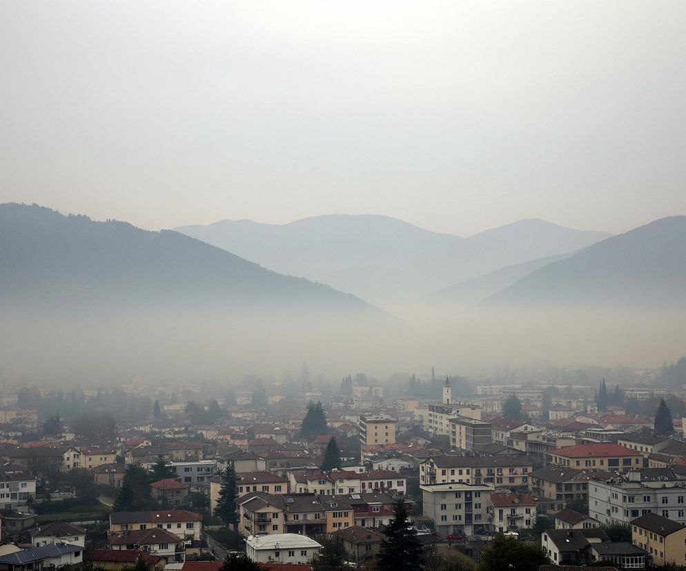 Panorama miasta położonego w dolinie, otoczonego przez góry, z widoczną gęstą mgłą lub zamgleniem. Na pierwszym planie widać liczne budynki mieszkalne i komercyjne o zróżnicowanej architekturze i kolorach dachów, dominują odcienie czerwieni, brązu i szarości. W tle wznoszą się zalesione stoki gór, których kontury stopniowo zanikają w mgle, a dalsze pasma górskie są prawie niewidoczne, zlewając się z jasnym, szarym niebem. Całość obrazu utrzymana jest w stonowanych, przygaszonych barwach, co potęguje wrażenie mglistej atmosfery.