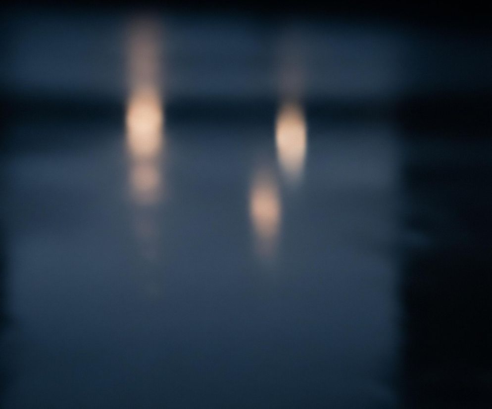 Ciemne, rozmyte tło w odcieniach granatu i czerni. W centrum widoczne są trzy pionowe, wydłużone plamy światła o ciepłym, pomarańczowo-beżowym zabarwieniu, z których dwie górne są wyraźniejsze, a jedna dolna mniejsza i bardziej rozproszona. Całość jest nieostra, tworząc abstrakcyjną kompozycję z efektem bokeh.