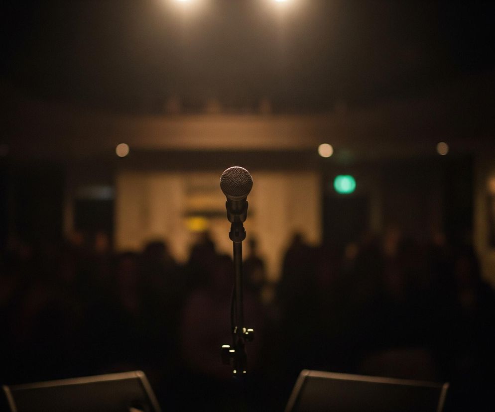 Mikrofon na statywie znajduje się na środku kadru, jego górna część jest jasno oświetlona, co tworzy wyraźny kontrast z ciemnym tłem. Za mikrofonem widoczne są rozmyte sylwetki ludzi oraz jasne punkty świateł, sugerujące, że znajdują się oni w audytorium lub na widowni. Dwa rozmazane, ciemne obiekty przypominające monitory sceniczne leżą na podłodze przed mikrofonem. Całość ma ciepłą, dominującą barwę brązu i czerni z kilkoma jasnymi punktami świetlnymi.