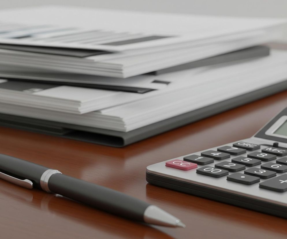 Na drewnianym, brązowym blacie biurka leżą obiekty biurowe. W centralnym punkcie znajduje się czarny kalkulator z zielonym wyświetlaczem, pokazującym szereg cyfr i czarnymi, kwadratowymi przyciskami z białymi napisami. Obok kalkulatora, po lewej stronie, leży czarny długopis z metalicznymi, srebrnymi elementami. W tle, częściowo rozmyte, ułożony jest stos białych dokumentów lub segregatorów.