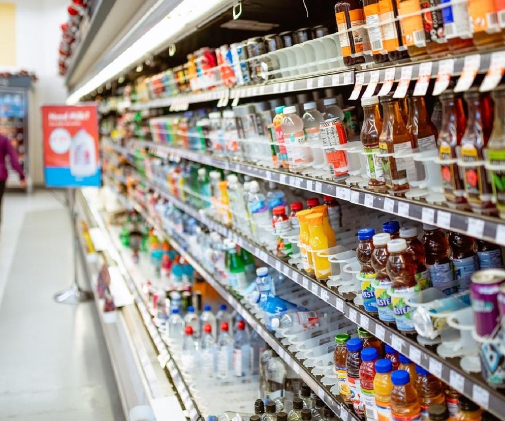 Półki w supermarkecie z napojami i artykułami spożywczymi, w tle rozmazana postać klienta. Obraz symbolizuje spadek cen żywności w Polsce, co analizuje Super Biznes.