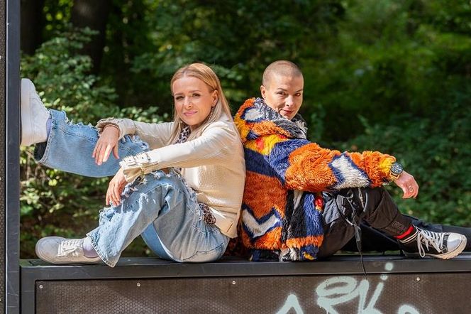 Aktorki Aneta Zając (Dominika) i Monika Dawidziuk (Karma) w luźnych stylizacjach, siedzące plecami do siebie na tle zieleni, promują odcinek 4097 „Pierwszej miłości”, gdzie ich bohaterki planują kradzież obrazu Edwarda, o czym przeczytasz na Super Seriale.