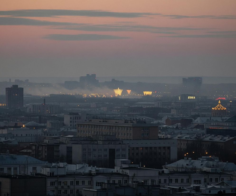 Panoramę miasta o zmierzchu lub świcie, zdominowaną przez szarość i pastelowe odcienie nieba. Na pierwszym planie widać gęsto zabudowane dachy budynków, niektóre z nich posiadają płaskie, szare powierzchnie, a inne pokryte są białym śniegiem lub lodem. W głębi obrazu, za miejską zabudową, rozświetlają się pojedyncze światła tworzące wzory, przypominające fajerwerki lub iluminacje, a dym lub mgła unosi się ponad nimi. Horyzont jest rozmyty, a nad nim rozciąga się niebo w kolorach od jasnoróżowego do fioletowo-szarego, z kilkoma ciemniejszymi chmurami.