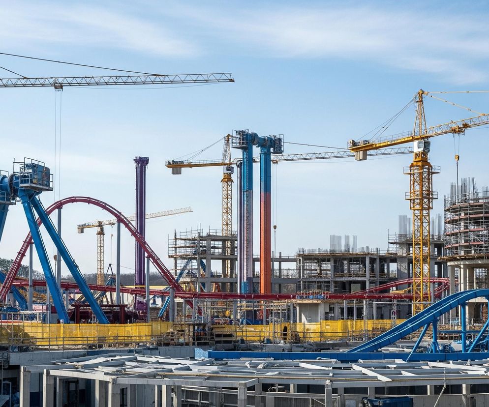 Rozległy plac budowy pod jasnym, błękitnym niebem z pojedynczymi smugami chmur. Dominują tutaj liczne, żółte żurawie budowlane o kratownicowych wysięgnikach, rozmieszczone w różnych częściach terenu. W centrum kadru znajdują się konstrukcje przypominające części lunaparku lub kolejki górskiej, z czerwonymi, zakręconymi torami i masywnymi niebieskimi, fioletowymi oraz pomarańczowymi wspornikami. Wokół nich wznoszone są betonowe budynki, niektóre o okrągłym kształcie, z widocznymi zbrojeniami i rusztowaniami. Na pierwszym planie widoczne są fragmenty niższych kondygnacji budynków, z szarymi, płaskimi elementami konstrukcyjnymi oraz żółtymi siatkami zabezpieczającymi na barierkach.