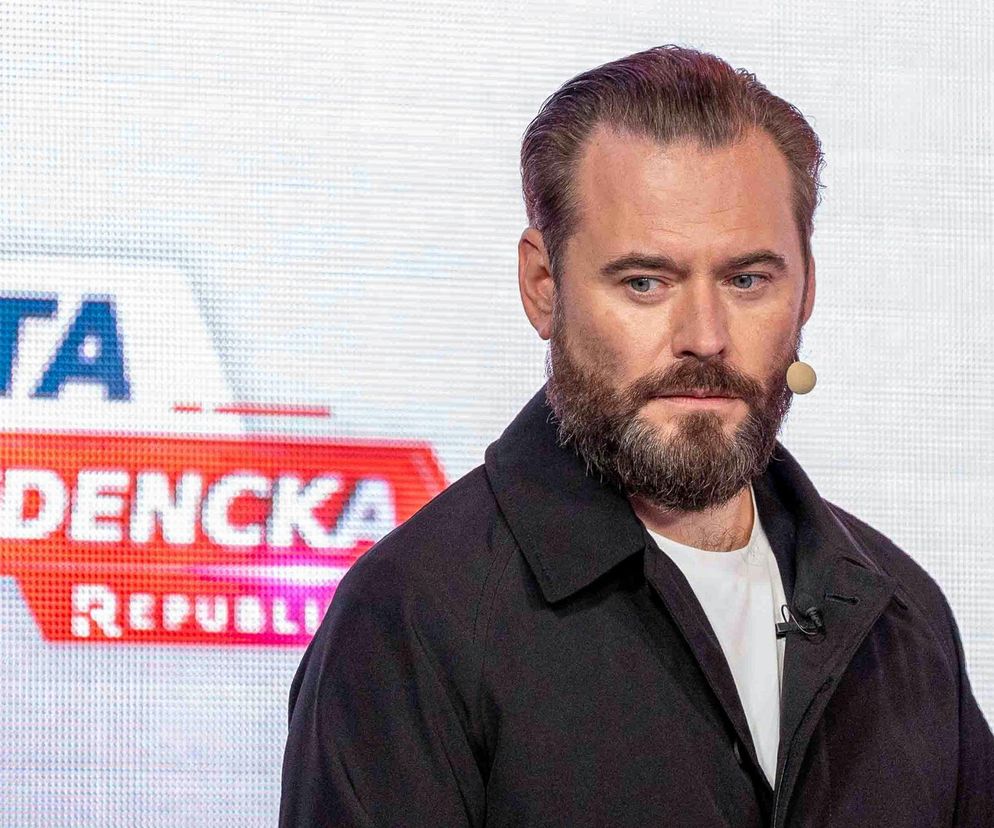 Krzysztof Stanowski z Kanału Zero, ubrany w czarną kurtkę i biały T-shirt, z charakterystyczną brodą, patrzy w dół. Za nim widać fragment napisu ATA i ZYDENCKA REPUBLI na jasnym tle. To kolejna kontrowersyjna inicjatywa, o której można przeczytać na Super Biznes.