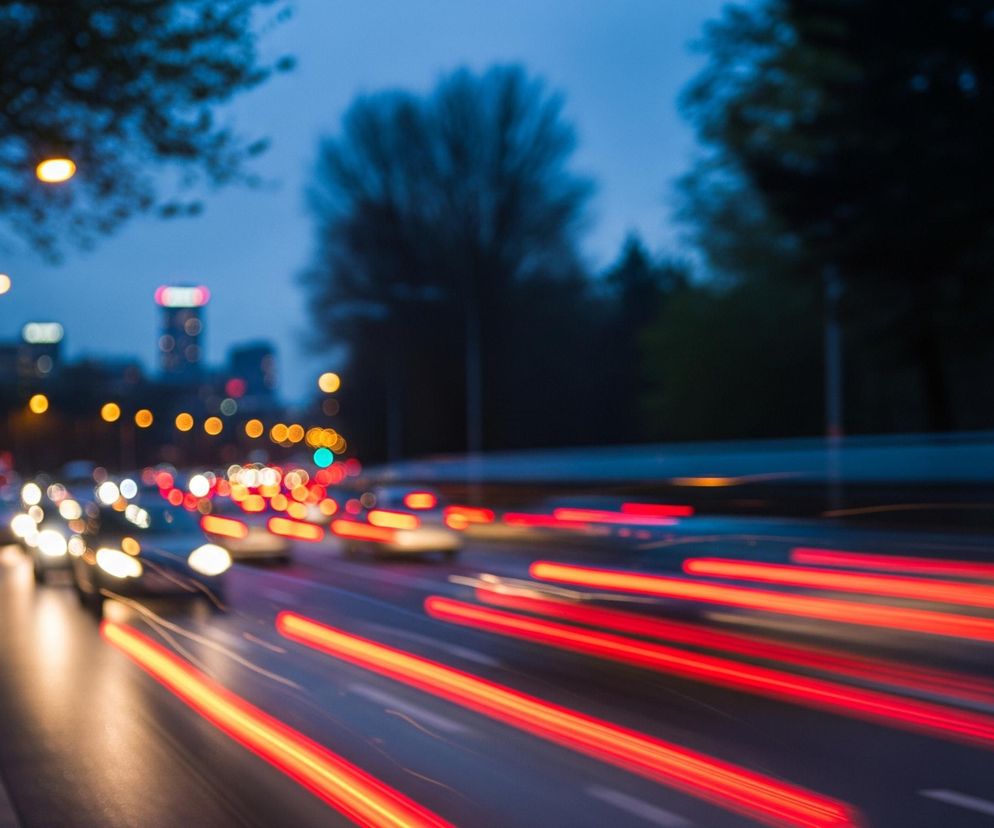 Nocna scena przedstawia miejską ulicę z rozmytym ruchem samochodów. Długa ekspozycja tworzy smugi światła: białe i żółte od reflektorów w kierunku patrzącego oraz długie, czerwone pasy od tylnych świateł pojazdów oddalających się w perspektywie. W tle widać niewyraźne, pomarańczowe punkty latarni ulicznych oraz zamazane światła budynków, a po lewej stronie ciemne kontury drzew. Całość utrzymana jest w ciemnych, chłodnych barwach, z dominującym granatowym niebem i odcieniami czerni i szarości na drodze i poboczu.