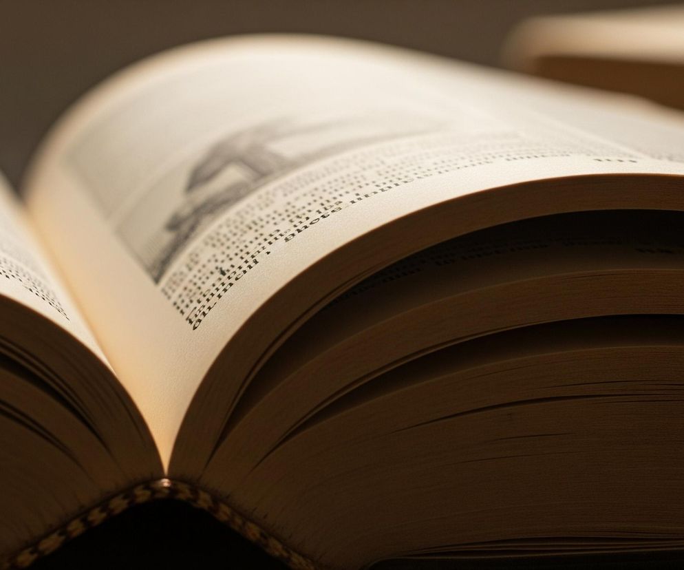 Otwarta książka leży na ciemnej powierzchni, ukazując swoje wnętrze. Strony, lekko pożółkłe od czasu, są pokryte drobnym, czarnym drukiem, a na lewej stronie widoczna jest również ilustracja. Grzbiet książki jest wyraźnie widoczny na dole zdjęcia, z widocznymi zszyciami lub ozdobnymi elementami. Światło pada z góry, oświetlając centralną część rozłożonych stron i tworząc delikatne cienie w fałdach kartek.