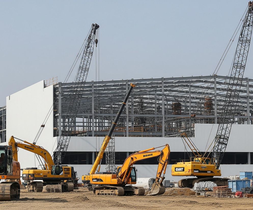 Plac budowy z dużą, niedokończoną konstrukcją budynku w tle i ciężkim sprzętem budowlanym na pierwszym planie. Budynek ma częściowo wzniesioną białą fasadę i widoczną metalową konstrukcję szkieletową na dachu. Na placu budowy znajdują się cztery duże, pomarańczowo-żółte koparki gąsienicowe oraz dwa czarne dźwigi kratowe, wszystkie w stanie spoczynku. Piaszczysty grunt jest nierówny, z widocznymi hałdami ziemi i różnymi materiałami budowlanymi, w tym niebieskimi kontenerami i drewnianymi paletami.
