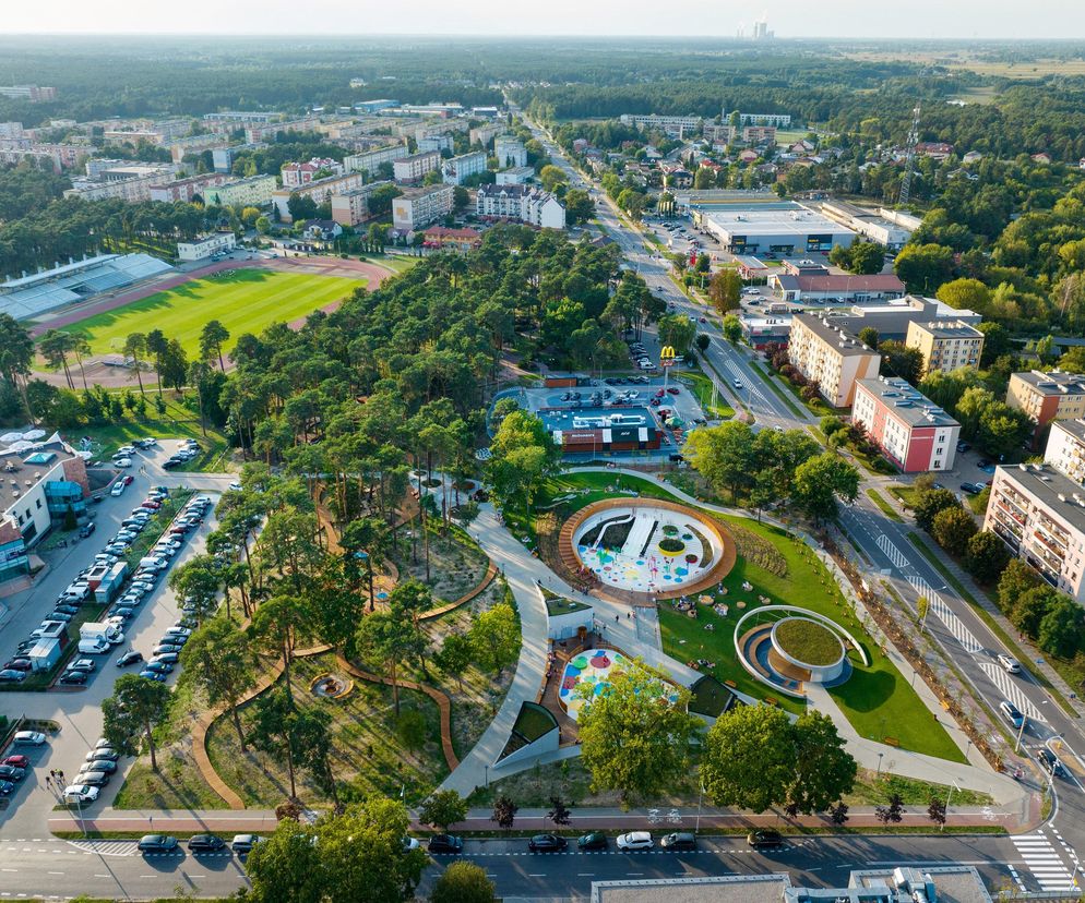 Park rekreacyjno-sportowy z wodnymi placami zabaw w Kozienicach autorstwa RS+ Robert Skitek, zgłoszony do konkursu ŻYCIE W ARCHITEKTURZE 2025. Widok z lotu ptaka ukazuje zielony teren z placami zabaw i ścieżkami wśród drzew, otoczony zabudową miejską, co jest przykładem udanej rewitalizacji przestrzeni publicznej, o której możesz przeczytać na portalu Architektura Murator Plus.