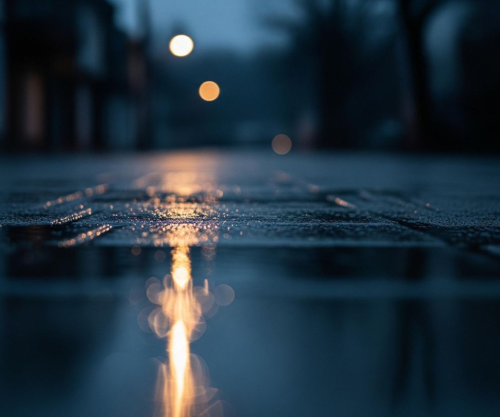 Droga z ciemnych płyt chodnikowych, pokryta drobnymi kroplami wody, odbija światła, tworząc na mokrej powierzchni długi, rozmyty pas jasnego, pomarańczowo-złotego blasku. W tle, rozmazane sylwetki budynków i drzew zlewają się z ciemnobłękitnym niebem, z którego wybijają się trzy rozmyte, okrągłe źródła światła, wyglądające jak latarnie uliczne. Głęboka perspektywa ukazuje ulicę uciekającą w dal.