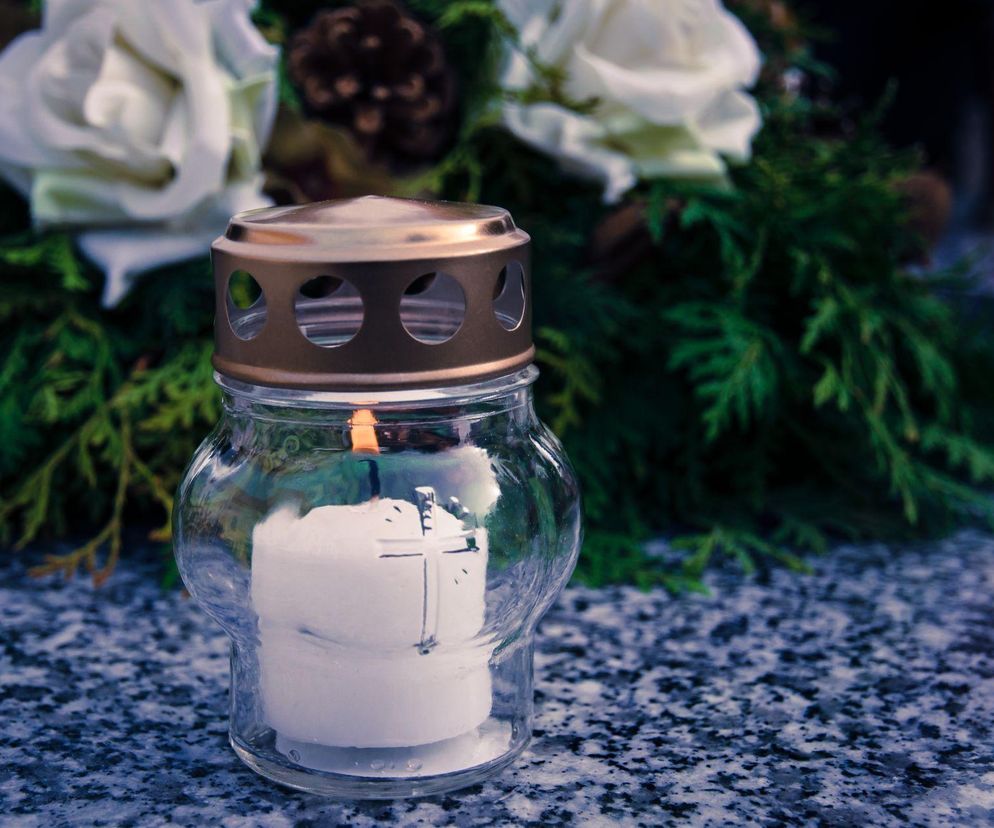 Szklany znicz z palącym się wkładem i metalową nakładką, z delikatnym krzyżem na wkładzie, stojący na granitowym nagrobku. W tle widać wieniec z białymi różami i zielonymi gałązkami. Więcej o trendach w branży zniczy przeczytasz na Super Biznes.