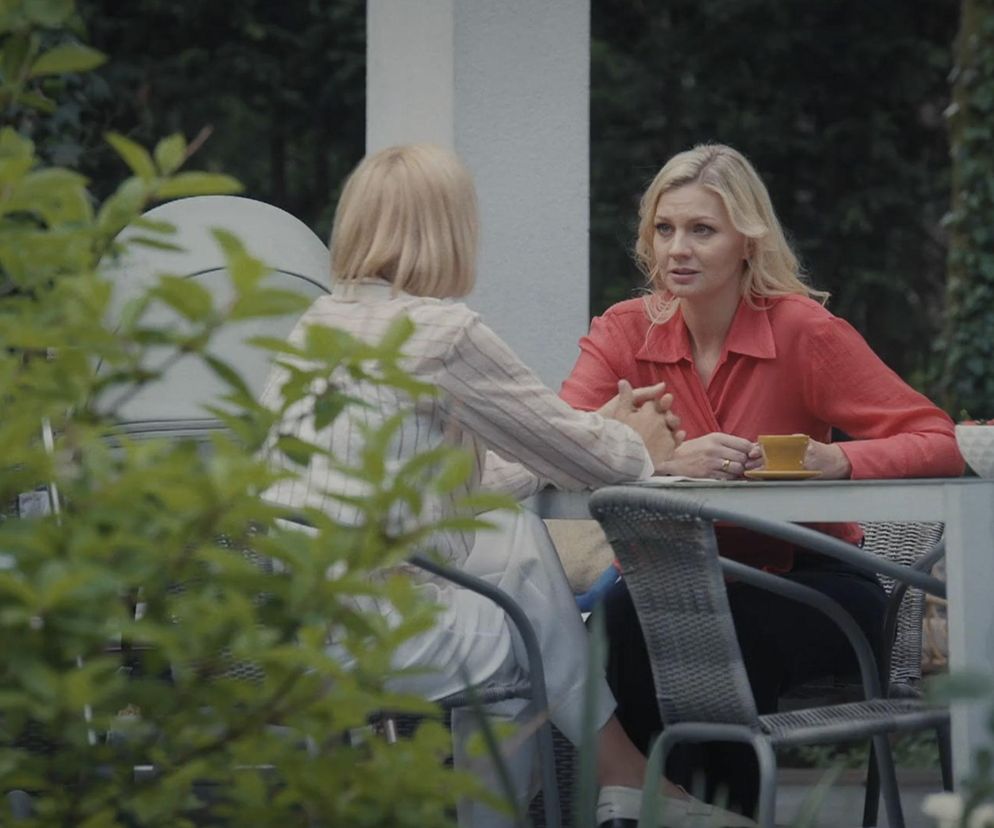 Dorota (Iwona Rejzner) i Jagoda (Katarzyna Kołeczek) siedzą przy stole w ogrodzie. Blondwłosa Dorota w czerwonej koszuli zamyślona opowiada coś blondwłosej Jagodzie, widocznej od tyłu. Scena z serialu M jak miłość dostępna na Super Seriale.