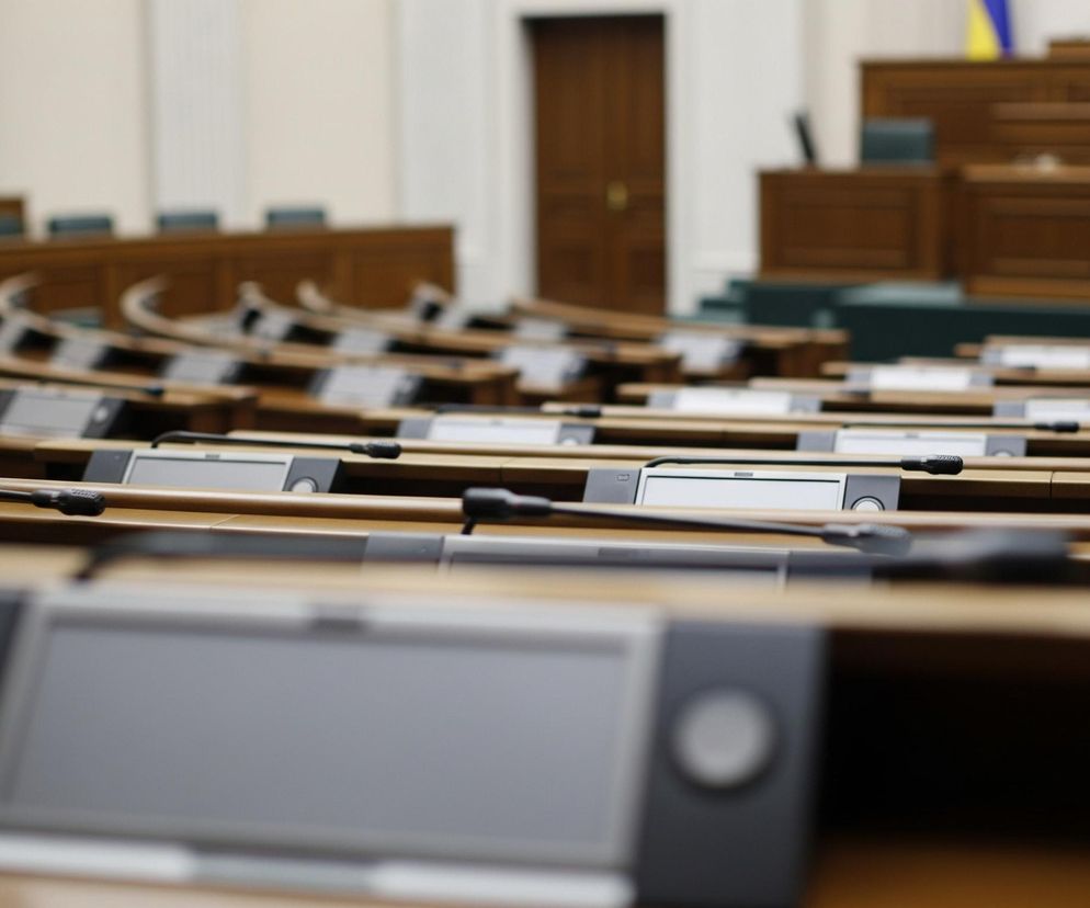 Puste, drewniane ławki z mikrofonami na wysięgnikach i panelami sterowania, rozmieszczone półkoliście, dominują w dolnej części kadru. Dalej w tle widoczne są kolejne rzędy identycznych miejsc, lekko rozmyte, wznoszące się schodkowo. Po prawej stronie, za rzędami ławek, znajdują się drewniane trybuny i fotele z ciemną tapicerką, a na tle ściany wyróżnia się pionowy fragment niebiesko-żółtej flagi.