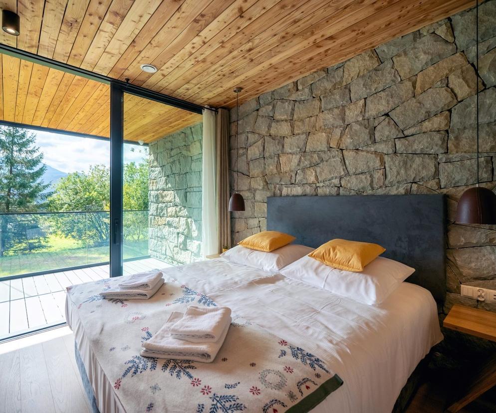 Sypialnia z kamienną ścianą i widokiem na góry. O projekcie Shelter House w Kościelisku przeczytasz na Architektura.