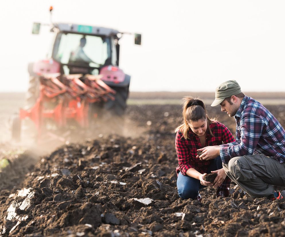Dwóch rolników, kobieta i mężczyzna, ogląda uprawianą glebę na polu, a w tle widać traktor z pługiem, co symbolizuje rolniczą pracę i wyzwania związane z dopłatami i nawozami. Więcej o ich problemach przeczytasz na Super Biznes.