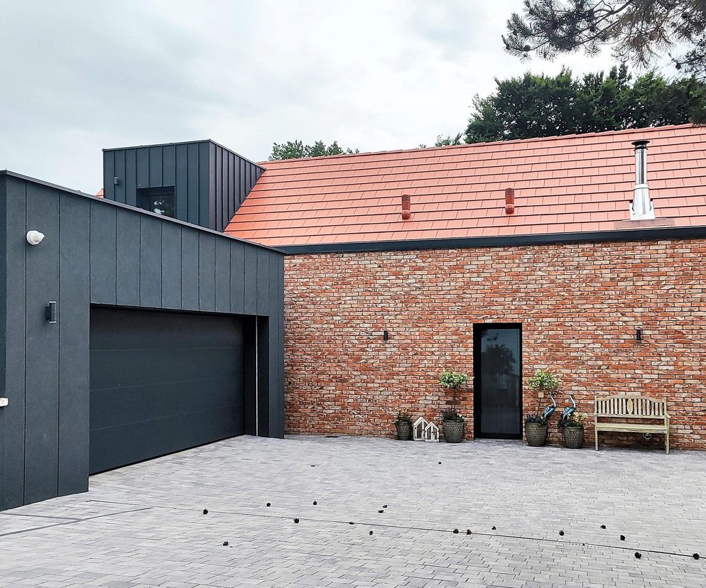 Frontowa część Domu nad Jeziorem z garażem o elewacji z ciemnych paneli i czerwoną ceglaną ścianą, oraz z bramą garażową, czarnymi drzwiami wejściowymi, ławką i donicami. To zgłoszenie do konkursu Życie w Architekturze 2025, o którym przeczytasz na Architektura Murator Plus.