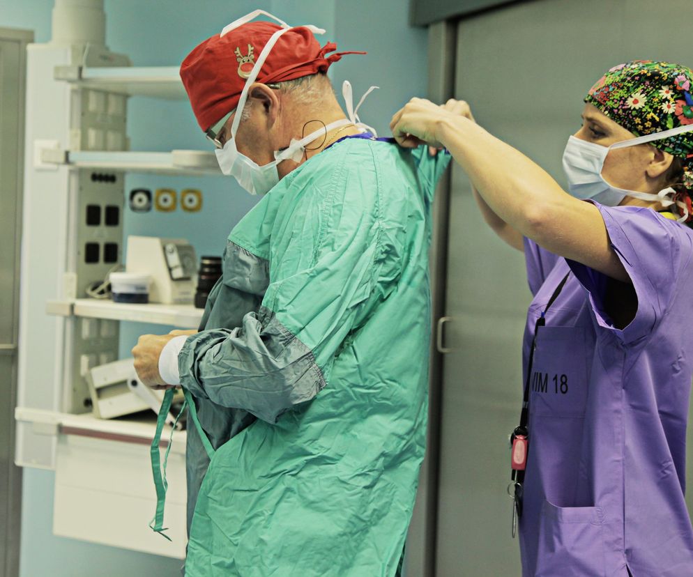 Para lekarzy przygotowujących się do operacji na sali operacyjnej. Lekarz w czepku z reniferem zakłada zielony fartuch, a pielęgniarka w czepku z kwiatowym wzorem pomaga mu go zawiązać. Scena przedstawia medyczne realia, o których przeczytasz więcej na Poradnik Zdrowie.