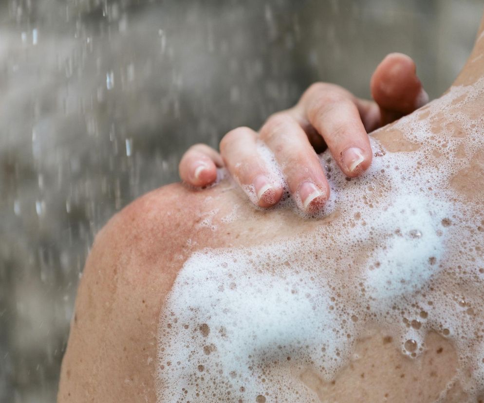Osoba pod prysznicem myjąca ramię, na skórze widać pianę z mydła i spływającą wodę, a jej dłoń delikatnie dotyka ramienia. Obraz symbolizuje codzienną higienę i jej znaczenie, o czym więcej przeczytasz na portalu Poradnik Zdrowie.